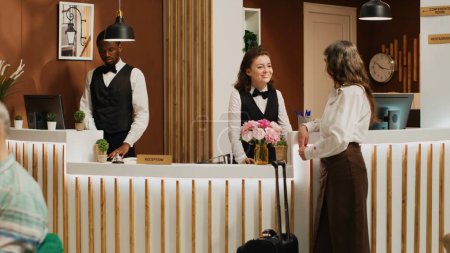 Téléchargez les photos : Une cliente s'enregistre dans le hall de l'hôtel de luxe avec l'aide d'un concierge amical. Réceptionniste saluant une femme âgée à la réception et aidant à l'inscription. Coup de main. - en image libre de droit