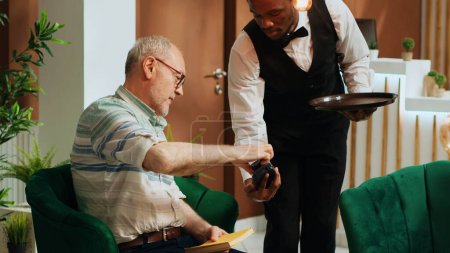 Foto de Personas mayores que pagan por la taza de café a través de tarjeta de crédito en el vestíbulo del hotel, pedir refresco del bar y hacer pos transacción. Bellhop sirviendo a invitado con bebida mientras él está esperando. - Imagen libre de derechos