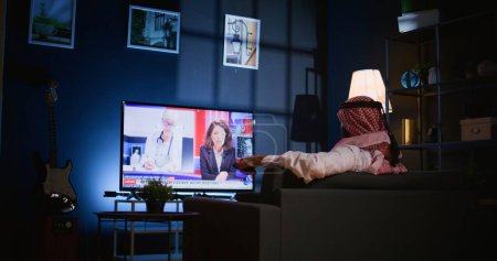 Arabischer Mann lümmelte auf Sofa und sah Nachrichtensendungen, während er es sich nach einem harten Arbeitstag im Wohnzimmer gemütlich machte. Nahost-Mensch bummelt auf Couch und schaut Fernsehkanal in schummriger Wohnung an