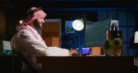 Lächelnder muslimischer Ingenieur, der mit Java-Programmiersprachen Skriptcode auf den Computerbildschirm schreibt. Selbstständiger Nahost-Entwickler arbeitet zu Hause an der Behebung von Datenbankfehlern