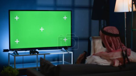 Arabischer Mann, der Nachrichten auf einem Chroma-Schlüsselgerät verfolgt, während er sich nach einem harten Arbeitstag zu Hause entspannt. Muslimische Person, die auf der Couch chillt, schaut auf vereinzelten Fernsehkanal