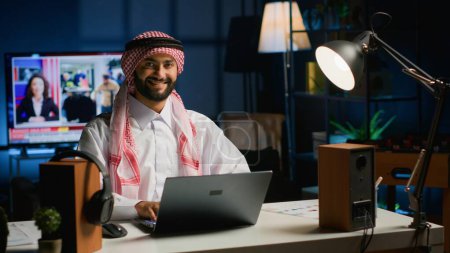 Téléchargez les photos : Portrait de tâches heureuses de résolution de pigiste arabe tout en travaillant à la maison, en tapant sur le clavier de l'ordinateur portable. Moyen-Orient homme navigation sur l'appareil numérique, la lecture des e-mails de clients dans le cadre de bureau - en image libre de droit