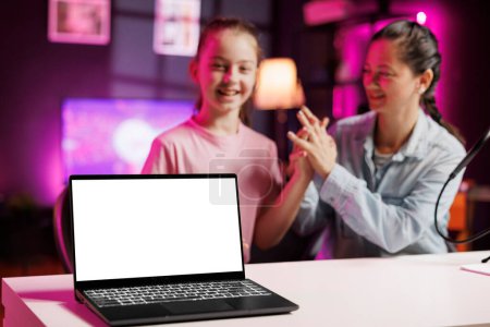 Cheerful girl next to her mother presents mockup laptop in pink neon lit living room used as vlogging studio. Young media star helped by parent to showcase isolated screen notebook device