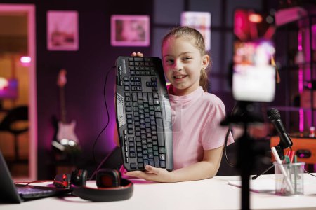 Talented cute girl enthusiastically presents latest gaming computer peripheral tech on her online channel, recording with phone on tripod. Young internet star filming wired mechanical keyboard review