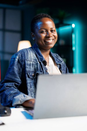 Retrato de blogger afroamericano trabaja en un portátil en un apartamento moderno. Smiling black woman es una freelancer dedicada, escribiendo y revisando información para su proyecto.