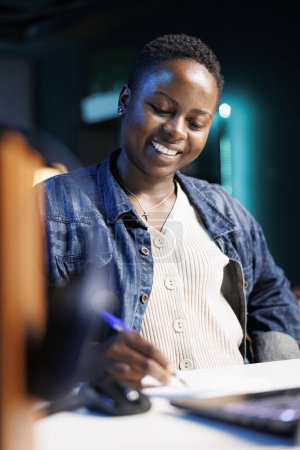Photo for Close-up of african american female blogger jotting down notes, thinking of new ideas for online content. Happy black woman sitting at desk, using pen and paper for studying and reviewing project. - Royalty Free Image