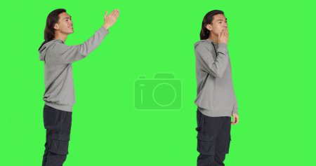 Asian guy sending air kisses on camera, acting romantic and cute standing against greenscreen background studio. Young adult feeling confident and joyful showing true honest emotions.
