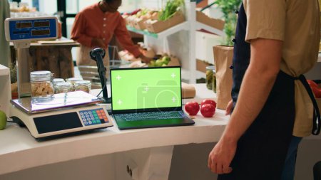 Shop owner works with greenscreen on laptop, sitting at register counter and waiting to serve customers in local zero waste supermarket. Vendor using blank display with chromakey mockup.