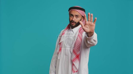 Foto de Hombre musulmán mostrando el símbolo de stop con la palma, presentando negatividad y desaprobación en el estudio. Joven expresando rechazo, descontento o desacuerdo sobre fondo azul. - Imagen libre de derechos