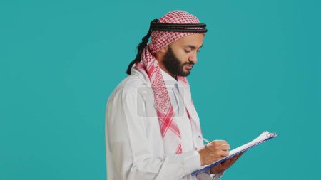 Foto de Hombre árabe escribiendo carta en papeles de pie sobre fondo azul, creando un nuevo horario y tomando notas. Adulto joven con ropa musulmana mirando la lista de verificación para el papeleo. - Imagen libre de derechos