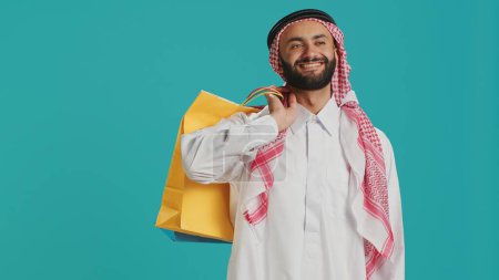 Muslim guy carrying many shopping bags in studio, posing with confidence after successful session of buying clothes and merchandise. Arab person purchasing products from mall on sale at retail store.