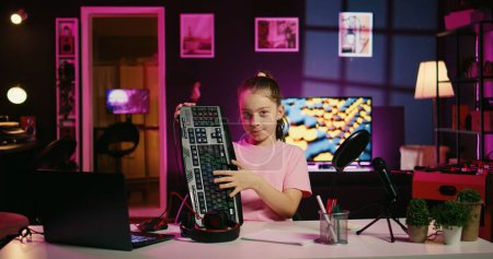 Child influencer filming keyboard, mouse and headphones review in home studio, showing capabilities and specifications. Little girl showcasing computer peripherals to fans on gen Z online channel