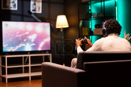 Mann spielt Science-Fiction-Singleplayer-Spiel auf Smart-TV, während er mit Freunden über Kopfhörer spricht. Spieler genießen Videospiel mit hochwertiger Grafik, mit Controller, um Raumschiff navigieren