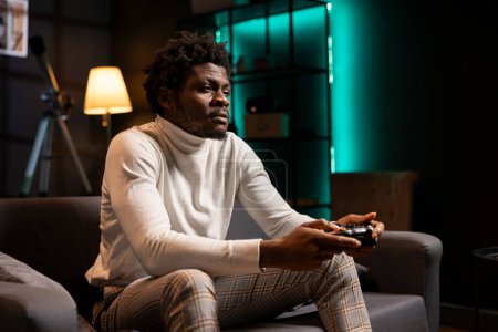 BIPOC player at home gaming and enjoying leisure time. Man sitting on couch in cyan neon illuminated living room, playing videogame on console, relaxing after hard day at work