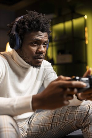 Téléchargez les photos : Un Afro-Américain jouant au jeu vidéo dans un appartement éclairé au néon jaune, tenant le contrôleur. Gamer participant au jeu multijoueur en ligne JcJ en utilisant le système de console et les écouteurs pour une expérience immersive - en image libre de droit