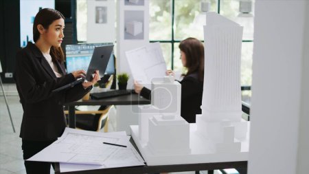 Asian contractor takes notes about 3d printed model on laptop, examining outline and scale of building maquette in architectural agency office. Cad specialist measuring layout for new project.