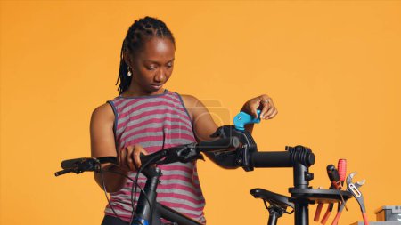 Mujer afroamericana instalando soporte de reparación de bicicletas, utilizándolo para ajustar desviador, la fijación de ruedas rotas, fondo de estudio. Profesional colocando bicicleta en el puesto de trabajo para hacer chequeo en él, cámara A