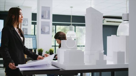 Architecture office employee looking at building model, working with printed project blueprints. Architect analyzing draft on house outline in coworking space, real estate agency worker.