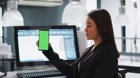 Developer shows mobile phone with greenscreen template, working in small business coworking space. Engineer using isolated copyspace layout on blank mockup screen, chromakey background.