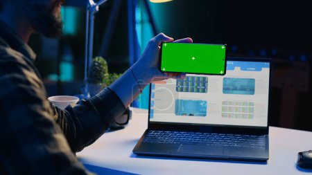 Photo for Man sitting on chair at home office desk in neon illuminated apartment holding green screen phone while working. Person in living room looking at business graphs on laptop and at mockup smartphone - Royalty Free Image