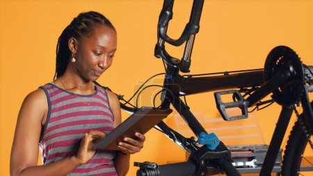African american woman doing bike servicing in studio background repair shop, looking on tablet maintenance list. Technician checking bicycle components that need repairing, camera A