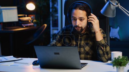Freelancer doing computer tasks for team project in personal home office while listening music. Remote employee working late at night, typing on laptop keyboard, putting headphones on, camera A