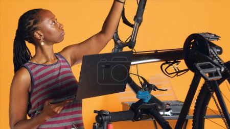 African american engineer doing bike servicing in studio background repair shop, looking on laptop maintenance list. Professional checking bicycle parts that need fixing, camera A