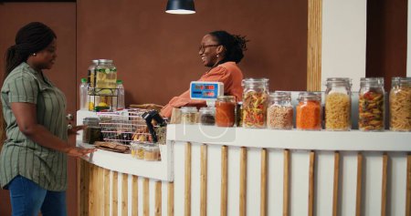 Mujer afroamericana que compra productos recién cosechados de la tienda de comestibles local, en busca de frutas y verduras maduras naturales. Mercader de ponderación de bienes a escala para el cliente. Disparo de mano.