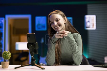 Cheerful girl excited to film with smartphone on tripod in living room used as professional studio for recording vlogs. Happy teenager discussing topics with fans, recording with phone