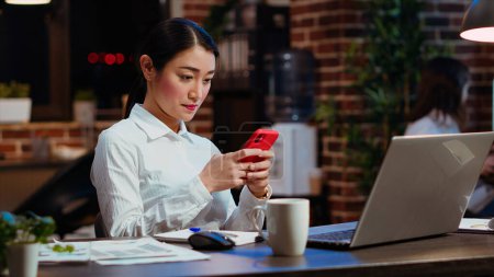 Photo for Smiling employee at work texting friends on cellphone, taking break from solving project tasks. Relaxed businesswoman using mobile phone to unwind in office late at night - Royalty Free Image