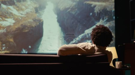 Foto de Hombre afroamericano viendo documentales de naturaleza con una impresionante cinematografía en una enorme pantalla de TV. Persona disfrutando de tomas de paisajes en televisor de pantalla ancha, relajarse en el sofá - Imagen libre de derechos