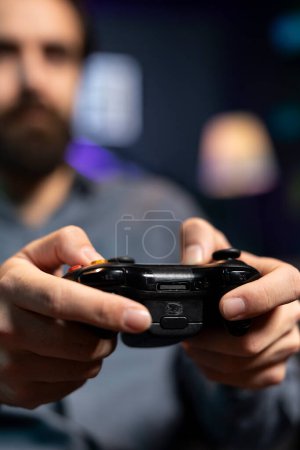 Focus shot on controller held by man defeating enemies while playing videogames on gaming console with teammates. Player in blurry background using gamepad to participate in esports game