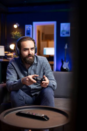 Gamer using headphones mic to discuss with teammates while playing games and enjoy leisure time. Excited man in apartment entertained by videogame on console, using gamepad