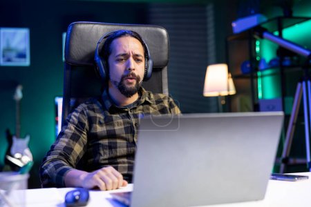 Photo for Male adult with headset is sitting at his desk immersed in a movie shown on his digital laptop. Young man wearing wireless headphones and watching a sad film on his portable computer at home. - Royalty Free Image