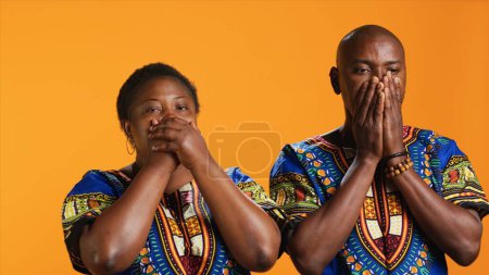 Ethnic couple doing three wise monkeys symbol on camera, showcasing sign to not hear, see or speak in studio. African american man and woman covering their eyes, mouth and ears.