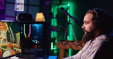 Hombre feliz usando micrófono de auriculares para comunicarse con amigos en línea desde su sala de estar oscura, jugar a videojuegos atractivos en PC de juegos en el escritorio de la computadora, relajarse después del trabajo