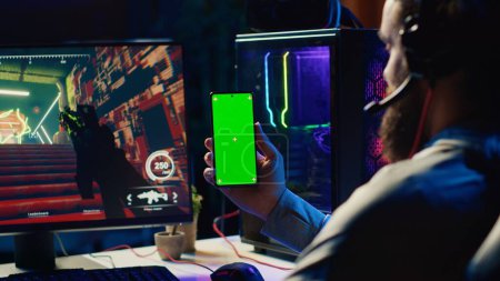 Man watching tutorial on green screen phone while playing first person shooter videogame with gun shooting laser bullets. Gamer learning to play game by looking at online guide on smartphone