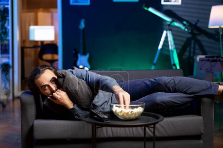 Persona abrazando almohada mientras atrapa series de televisión suspendidas en servicios de streaming basados en suscripción, disfrutando de palomitas de maíz. Hombre asustado en casa viendo programas de VOD de terror en la pantalla de televisión, comiendo bocadillo