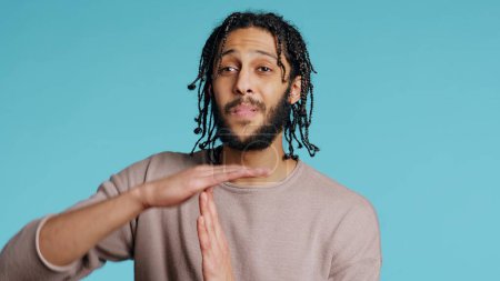 Assertive Middle Eastern man asking for timeout, doing hand gestures. BIPOC man doing pause sign gesturing, wishing for break, isolated over blue studio background, camera B