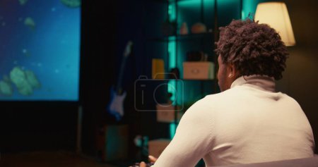 Téléchargez les photos : Homme afro-américain utilisant le service de jeu en nuage pour jouer exigeant jeu vidéo de science-fiction sur grand écran de télévision. Joueur jouissant de graphismes de haute qualité, jeu en streaming en ligne, zoom arrière - en image libre de droit