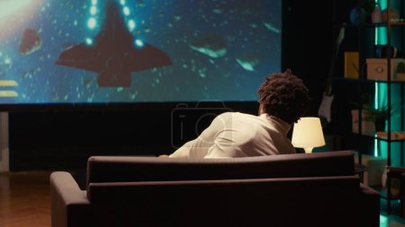 Téléchargez les photos : L'homme afro-américain joue au jeu solo de science-fiction avec un HUD superposé montrant des barres de santé et minimisant sur un énorme écran de télévision. Gamer jouissant de graphismes de traçage de rayons de haute qualité dans le jeu vidéo console - en image libre de droit