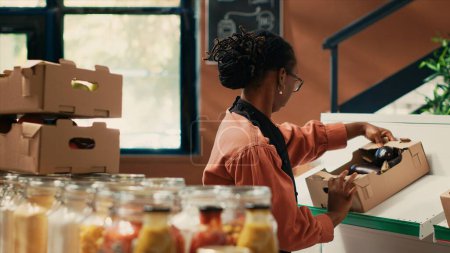 Comerciante organizando frutas y verduras en cajas en estantes, preparando mercancía fresca en el mercado de agricultores antes de abrir la tienda. Mujer afroamericana arreglando productos naturales.