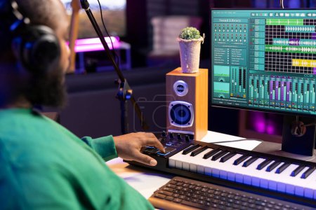 Music engineer adding synthesizer notes over his audio files in home studio, editing tracks with digital software and producing new songs for an album. Artist works with mixing console and gear.