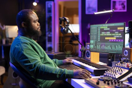 Artiste afro-américain composant une nouvelle bande son pour un projet, en utilisant un logiciel de production professionnel dans son home studio. Chanteur créatif compétent produisant de la musique et ajoutant des effets sonores.