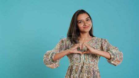 Portrait of jolly friendly indian woman doing heart symbol shape gesture with hands. Cheerful nurturing person showing love gesturing, isolated over studio background, camera A