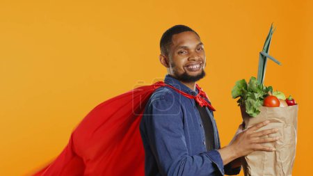 Young adult acting as a superhero with a red cape presenting homegrown produce in a paper bag, advocating for sustainable lifestyle concept. Person supporting organic food. Camera B.