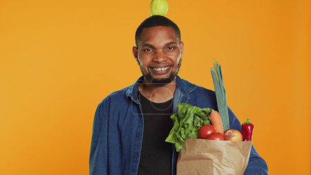 Young adult trying to keep balance with an apple on his head in studio, playing around and advocating for zero waste and sustainable lifestyle. Model carries paper bag with bio goods. Camera A.