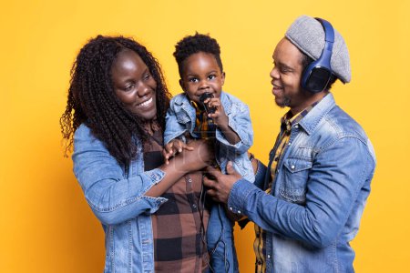 Glückliche Mama und Papa hören ihrem kleinen Kind im Studio zu, wie es ein Lied singt, der afrikanisch-amerikanische Vater hält sein Mikrofon in der Hand und trägt ein Headset. Süße Familie aus drei auftretenden Melodien.