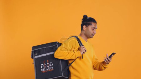 Man holding thermal backpack answering phone, fulfilling clients orders, studio backdrop. Food delivery service worker doing delivery to customers, talking on smartphone, camera B