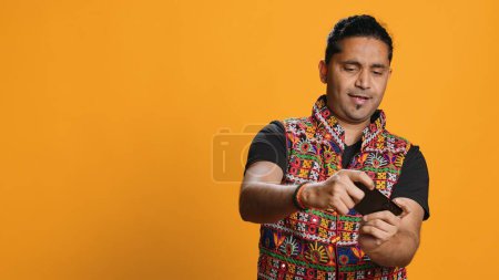 Happy Indian man entertained by videogames on smartphone, enjoying leisure time. Gamer enjoying game on phone, having fun defeating enemies, isolated over studio background, camera A